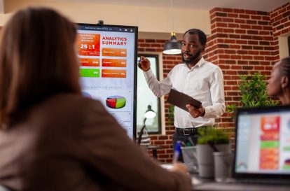 Businessman with clipboard in hand, using digital monitor to explain analytics summary in meeting. African american entrepreneur doing project presentation, planning marketing strategy with his team.